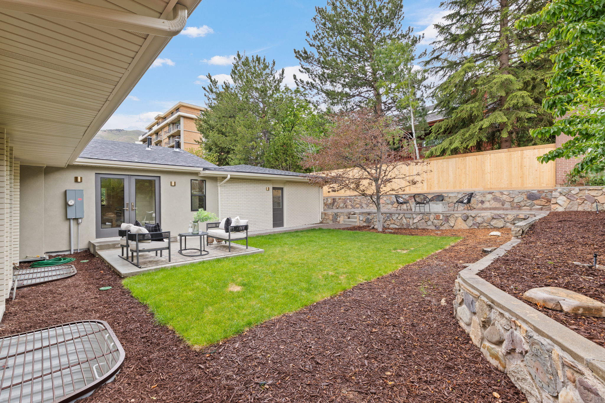 Private backyard featuring a concrete patio with French doors, manicured lawn, tiered landscaping with stone retaining walls, and a natural wood fence