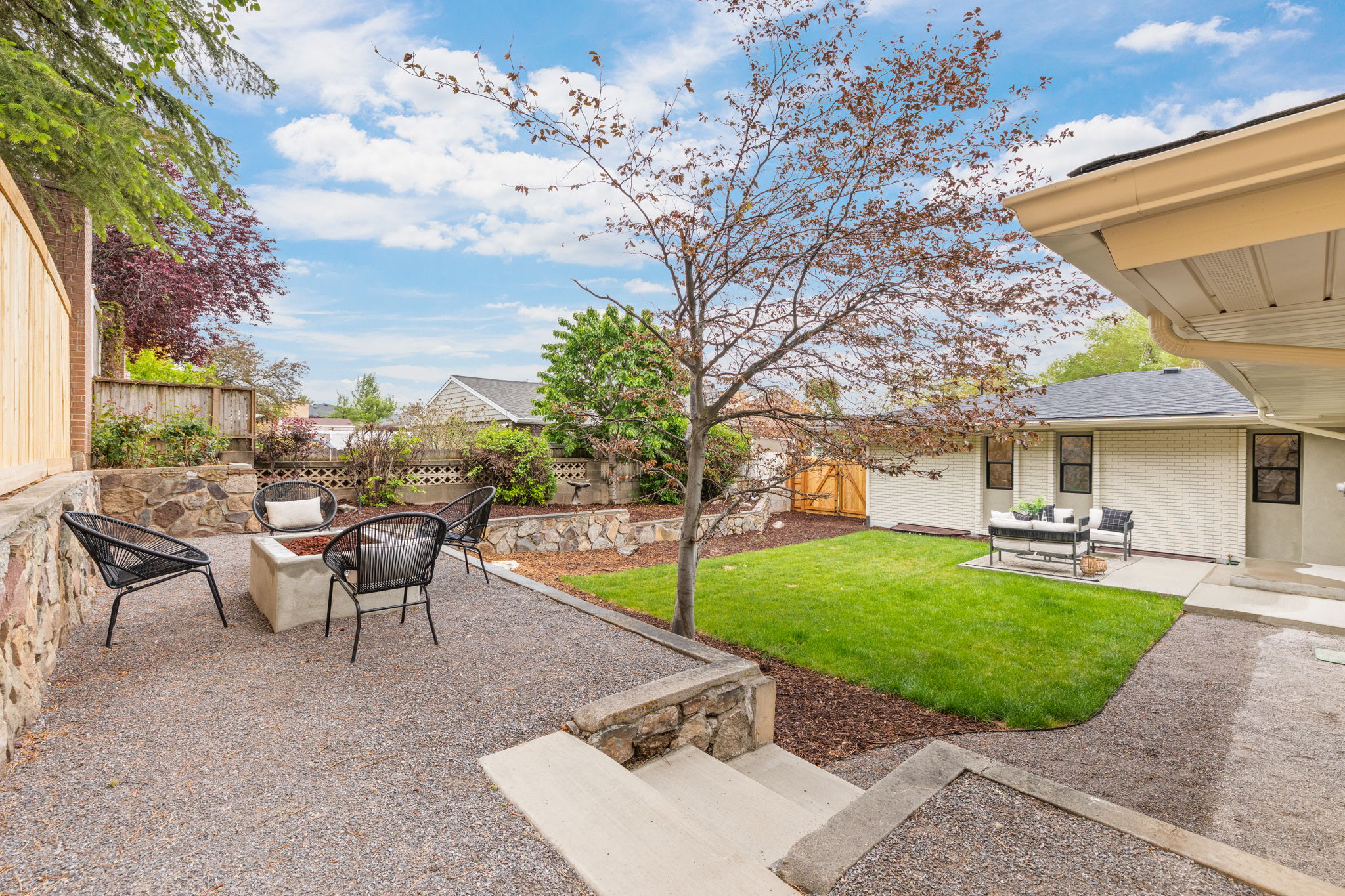 Spacious backyard featuring a gravel patio with a built-in fire pit, a manicured lawn, and mature landscaping