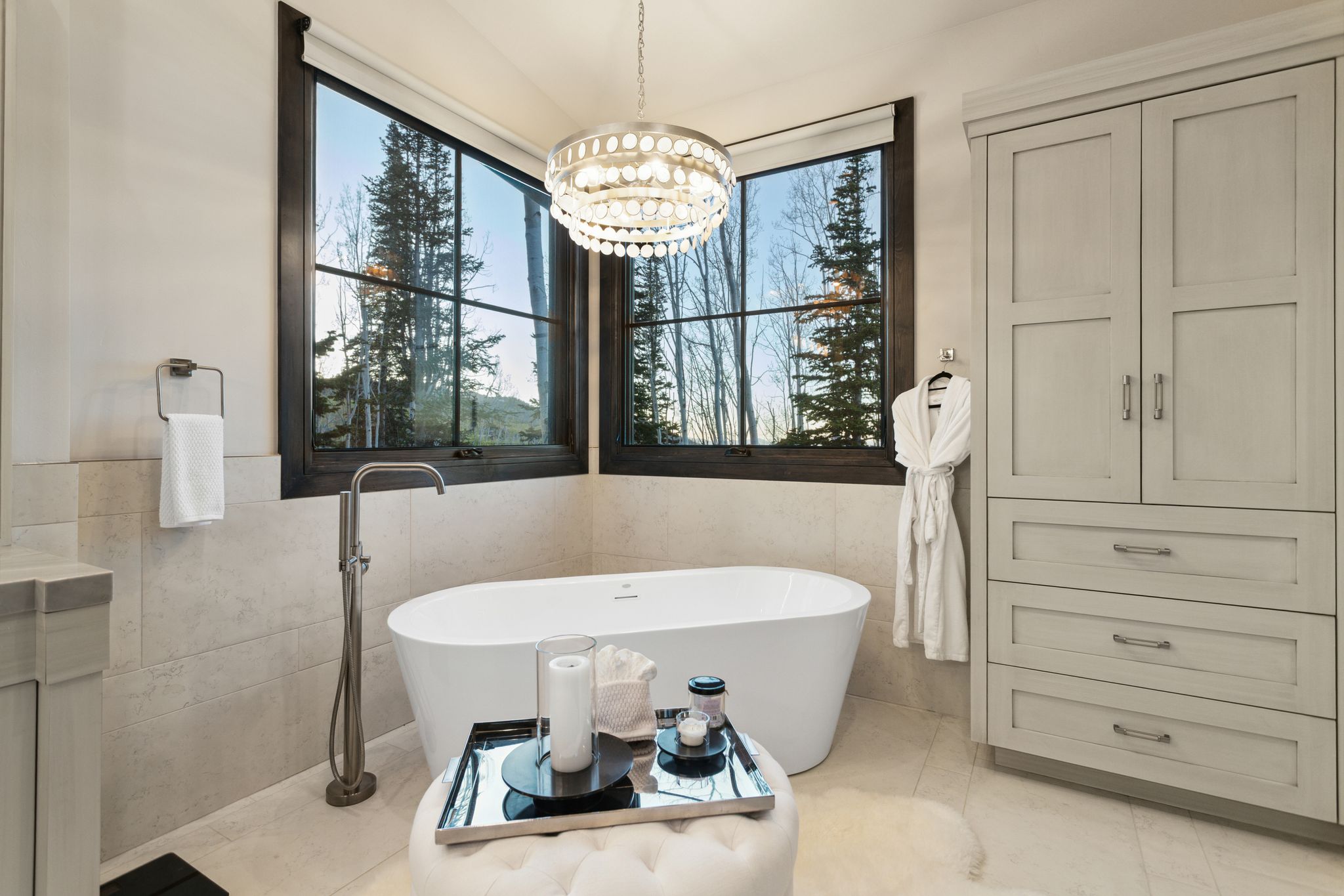 Freestanding soaking tub with a floor-mounted faucet, corner windows with dark frames, and a contemporary chandelier