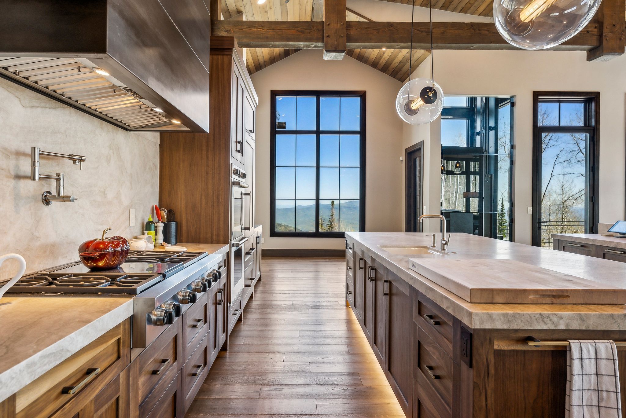 Gourmet kitchen featuring a professional range with pot filler, custom wood cabinetry, extensive natural stone countertops, a large center island with integrated sink, and vaulted ceilings with exposed wood beams