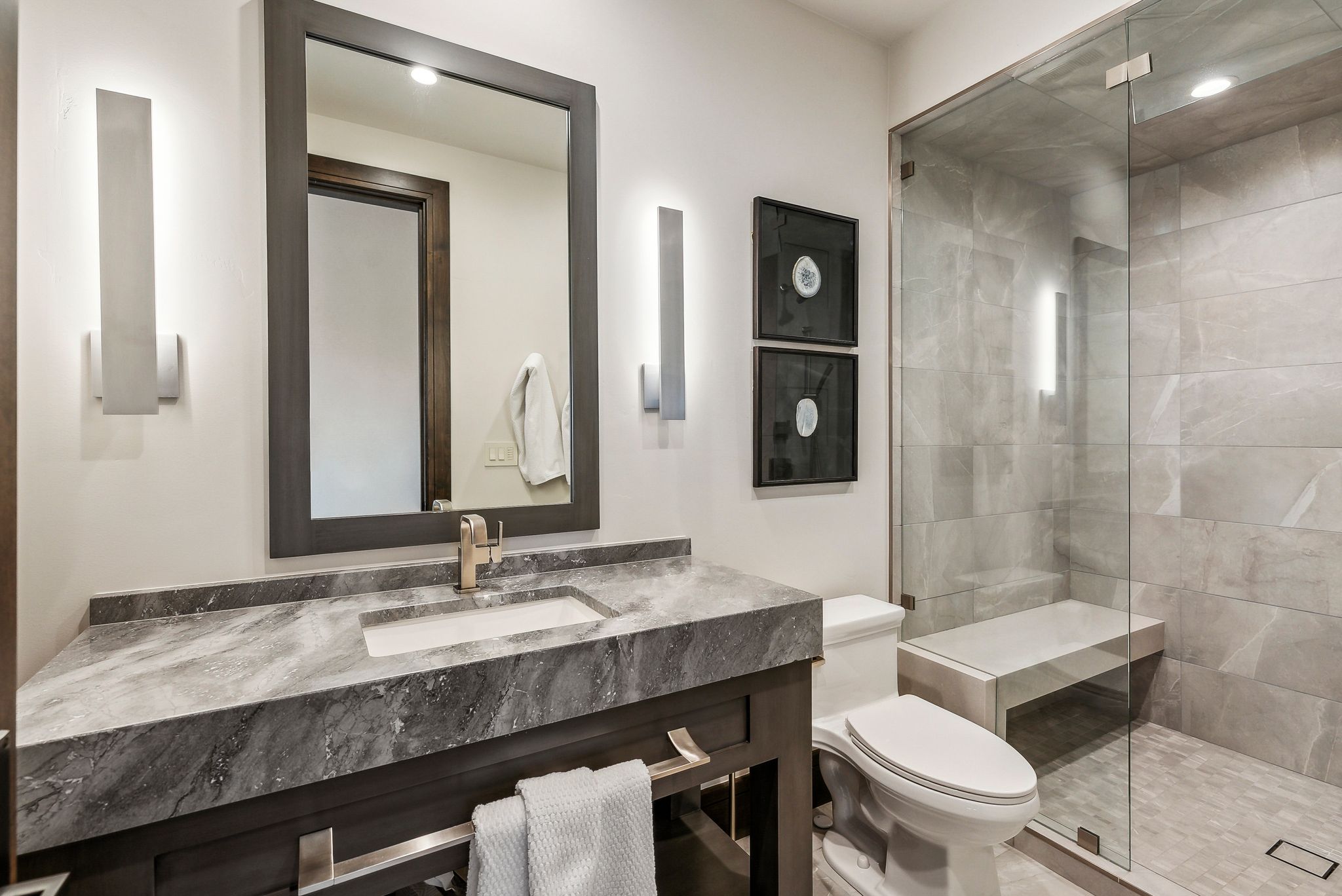 Bathroom featuring a dark wood-finish vanity with a stone countertop and integrated sink, a framed mirror, and contemporary sconce lighting