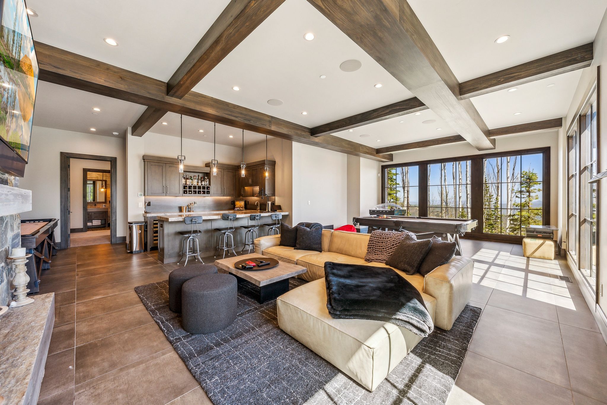 Spacious living area featuring exposed wood ceiling beams, a built-in wet bar with pendant lighting, a stone fireplace, large windows with exterior scenery, and durable tile flooring