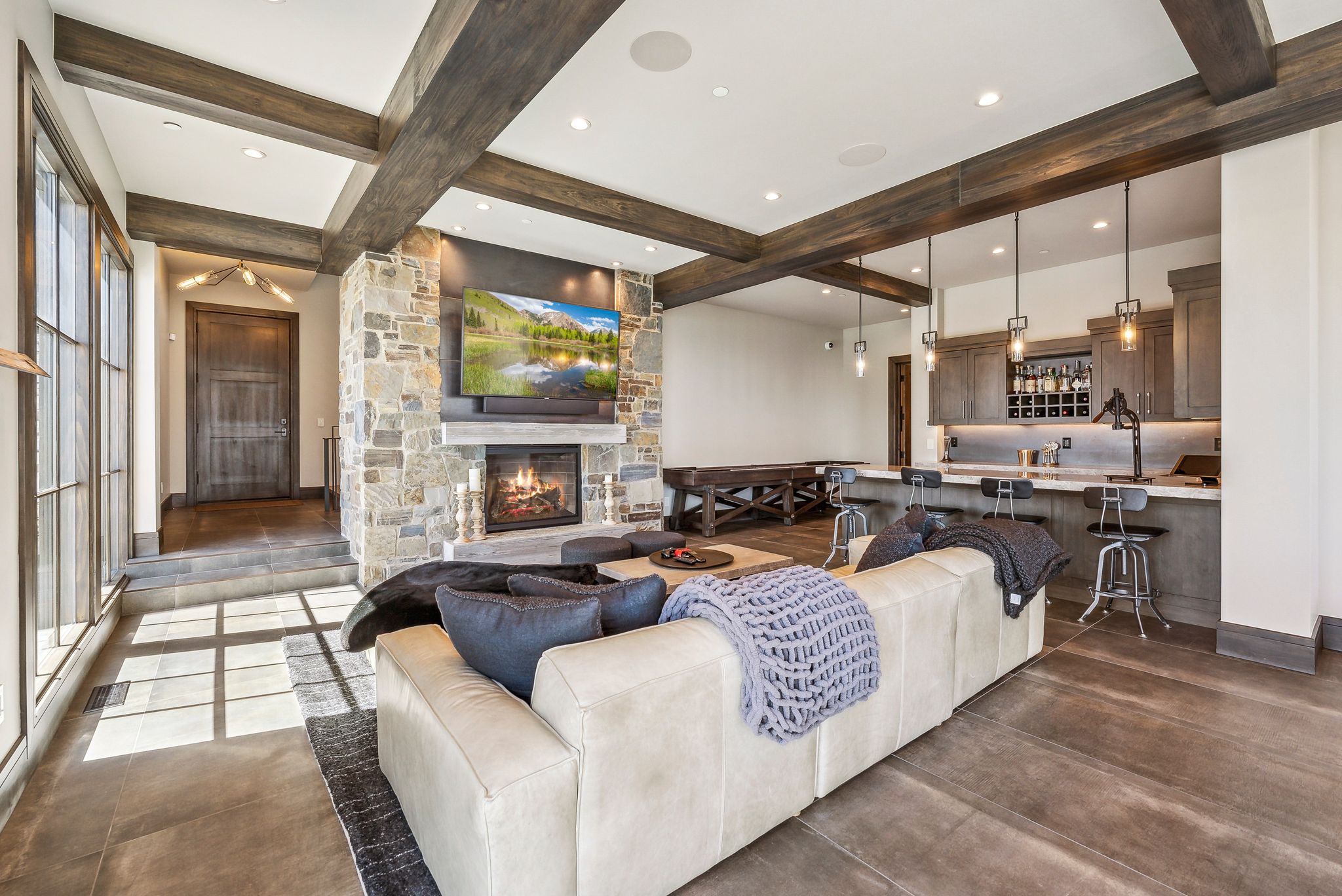Spacious living area featuring a stone-clad fireplace, wood-beamed ceiling, built-in wet bar with pendant lighting, and large windows
