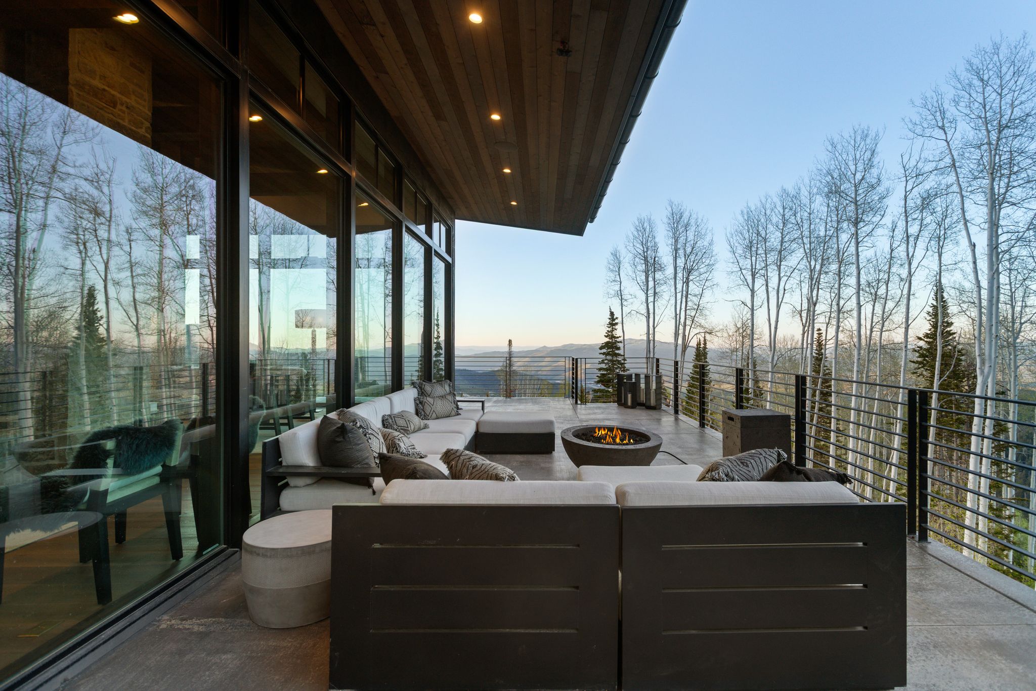 Expansive outdoor deck with a built-in fire pit, glass panel sliding doors, wood-paneled ceiling, and metal cable railings