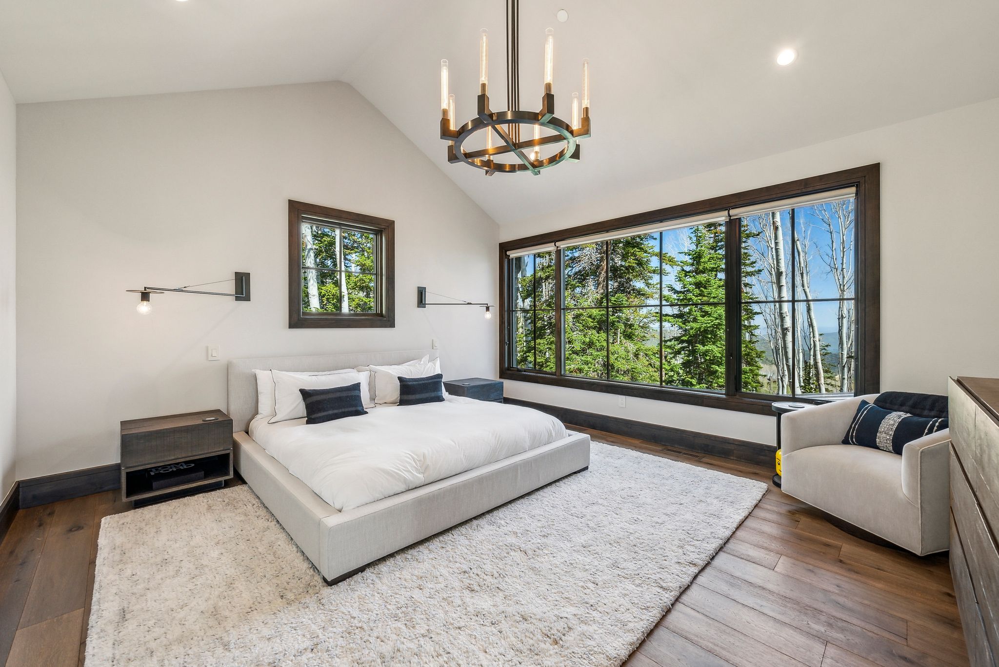 Spacious bedroom featuring vaulted ceilings, a large picture window with dark trim, rich wood-finish flooring, and a contemporary chandelier