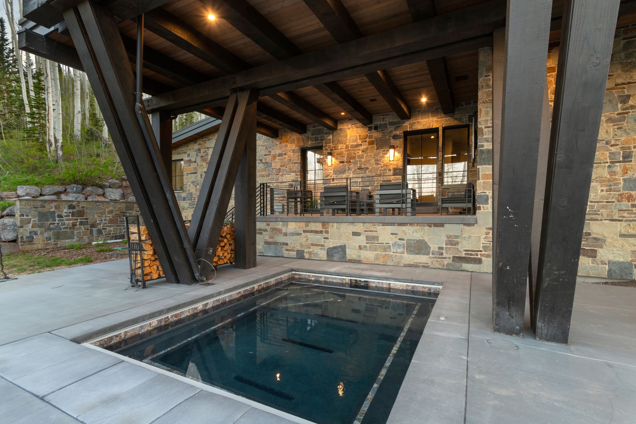 In-ground plunge pool surrounded by a concrete patio, featuring a stone exterior and a covered deck with exposed timber beams and recessed lighting