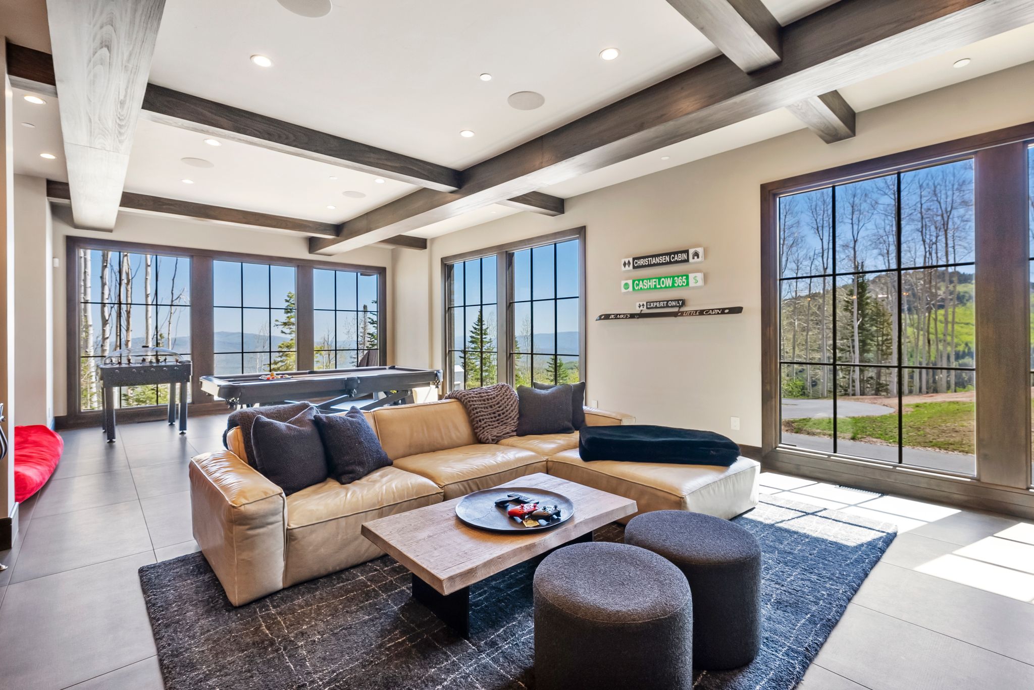 Expansive entertainment room featuring large grid windows, wood-finish ceiling beams, recessed lighting, and tile flooring