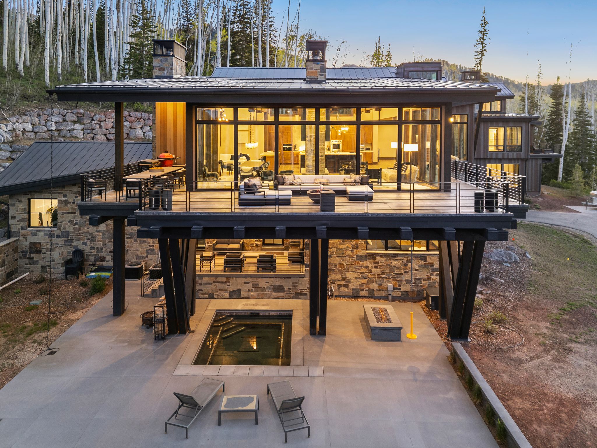 Modern architectural residence featuring multiple levels of stone and wood-paneling, a spacious deck with metal railings, an integrated spa, and a ground-level patio with a built-in fire pit