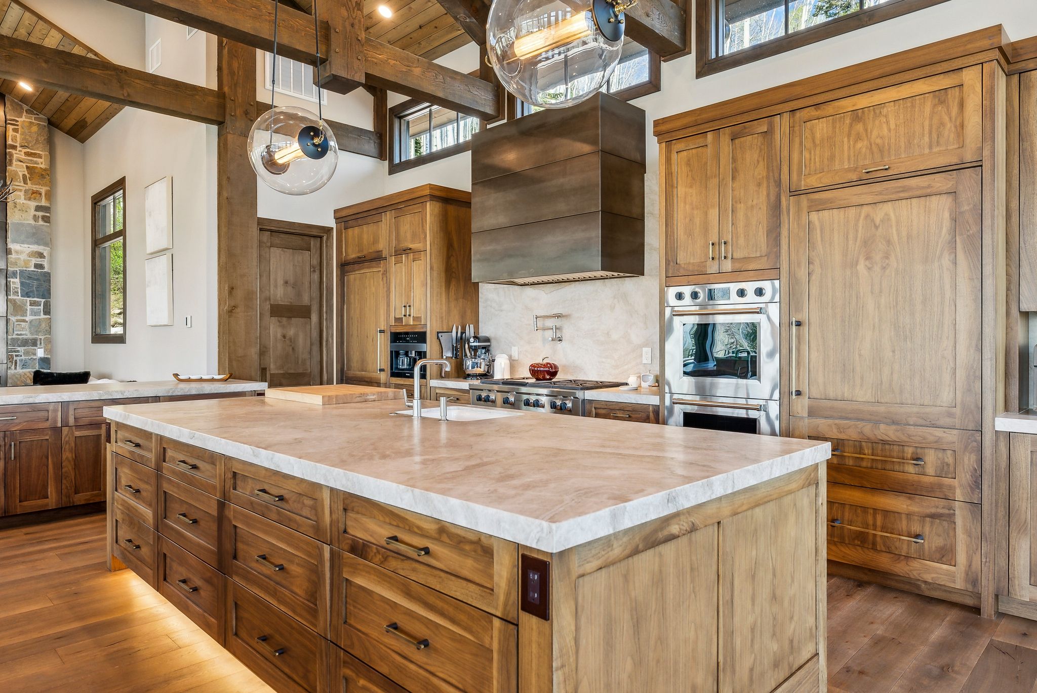 Spacious kitchen featuring a large central island with extensive storage, wood-finish flooring, exposed wooden beams, integrated stainless steel appliances, and a stone-clad accent wall