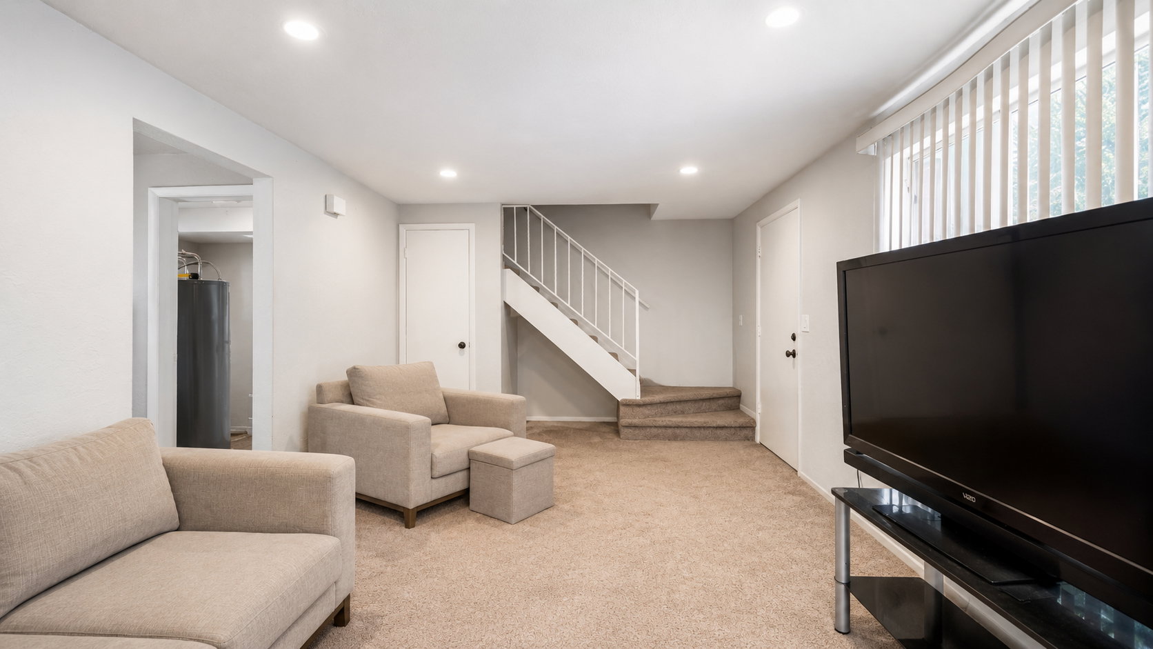 Spacious carpeted room featuring recessed lighting, a staircase with white railings, and a window with vertical blinds