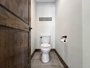 White toilet with a dark-finish flush lever, wood-finish flooring, dark wood baseboards, and a dark wood door with dark hardware