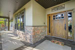 Covered front porch featuring stacked stone accents, multiple window panes, a solid wood entry door with glass inserts, and a coordinating wood-paneled ceiling