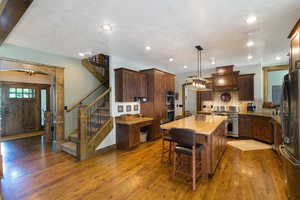 Open-concept kitchen and entryway featuring wood-finish flooring, a wood staircase with wrought iron balusters, a large kitchen island with counter seating, stainless steel appliances, and built-in wood cabinetry