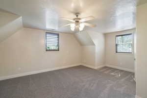 Spacious room featuring vaulted ceilings, a ceiling fan with integrated lighting, two windows for natural light, and plush gray carpeting