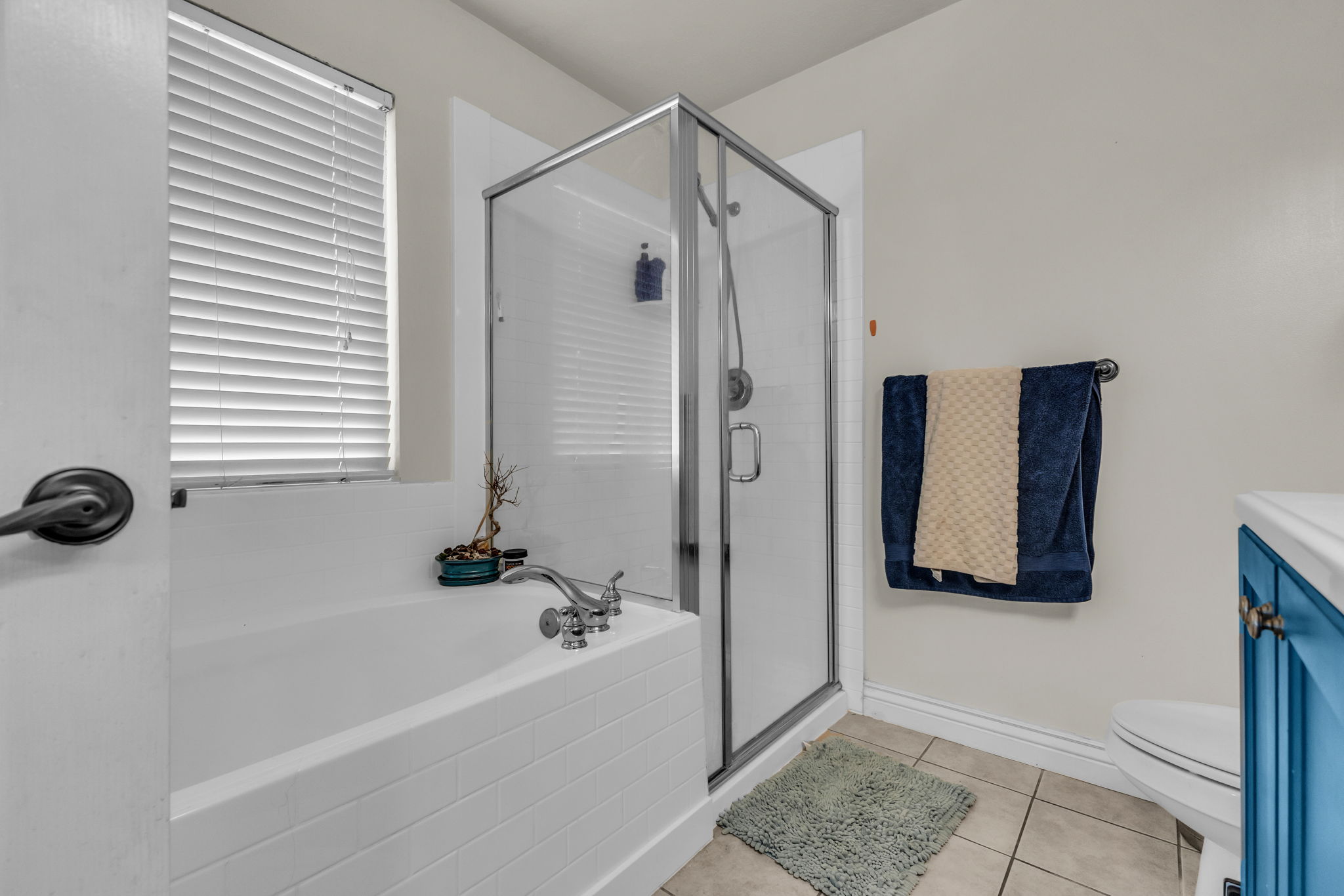 Bathroom featuring a white bathtub, a clear glass shower enclosure, beige floor tiles, and a window with white blinds