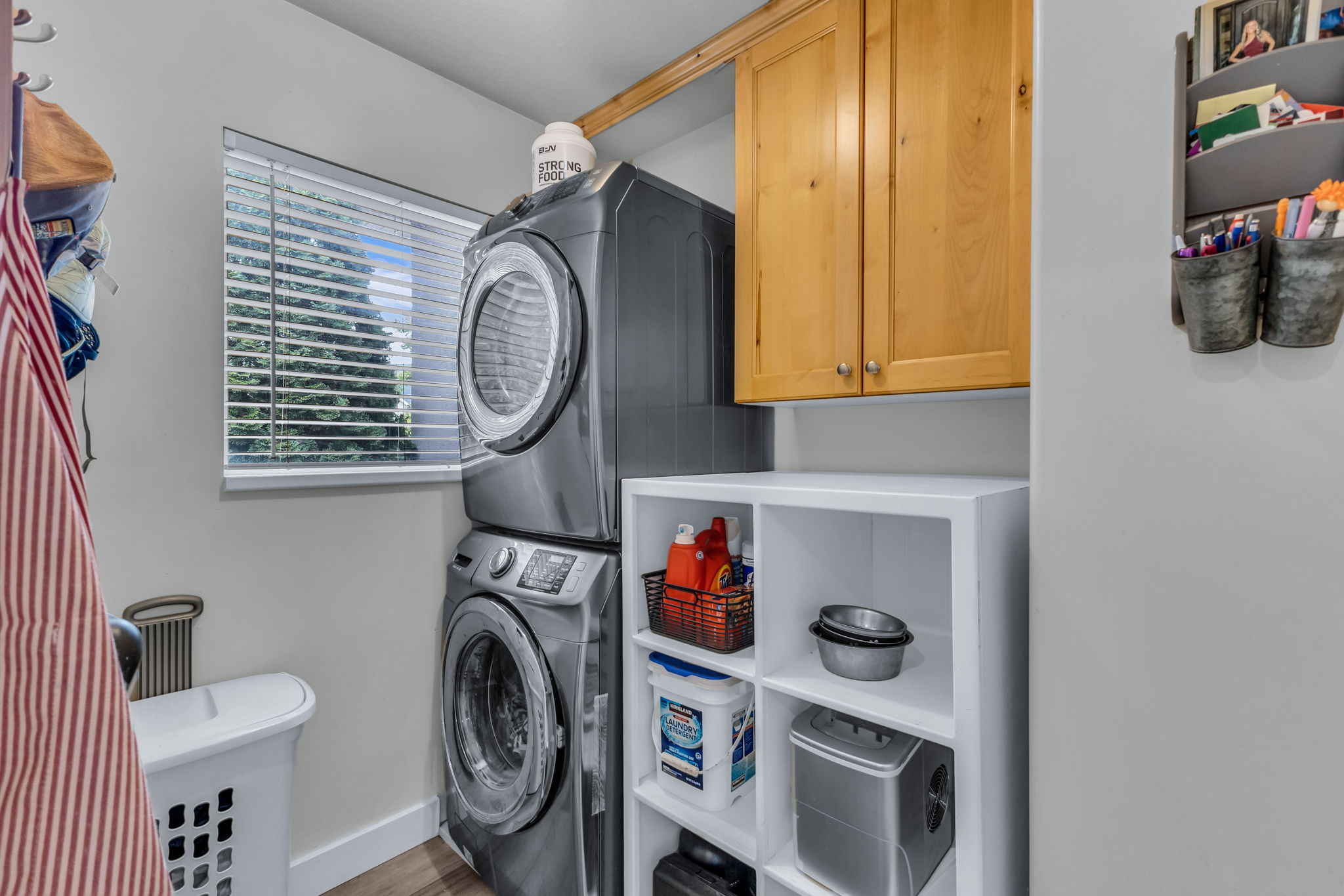 Dedicated laundry area featuring modern stacked appliances, a window with horizontal blinds, built-in natural wood cabinetry, and open shelving storage