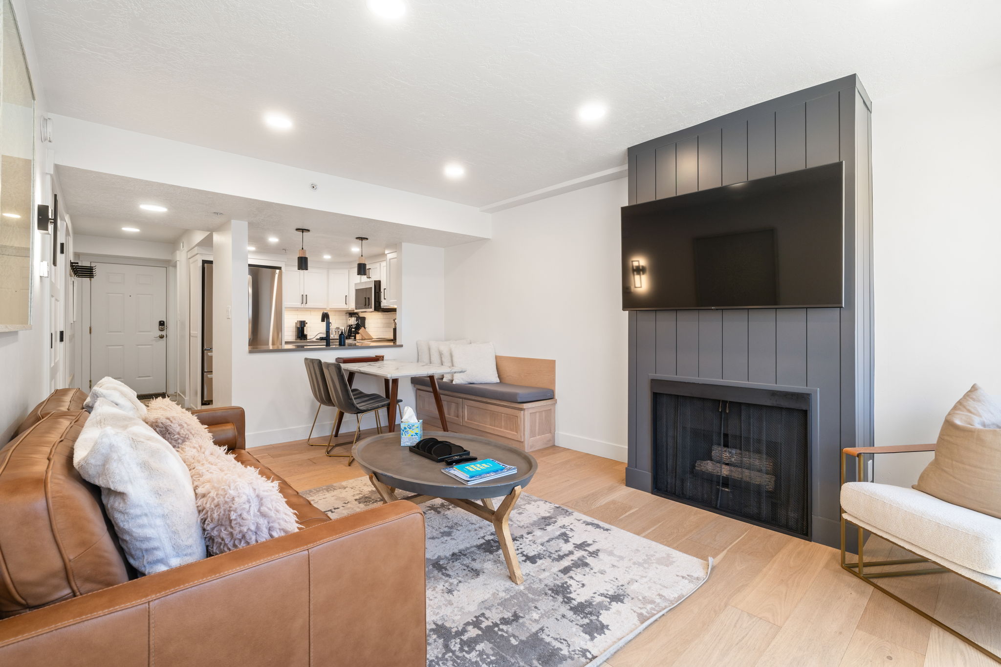 Living area featuring a modern fireplace with a dark paneled surround, light wood-finish flooring, recessed lighting, and a partial view of the kitchen