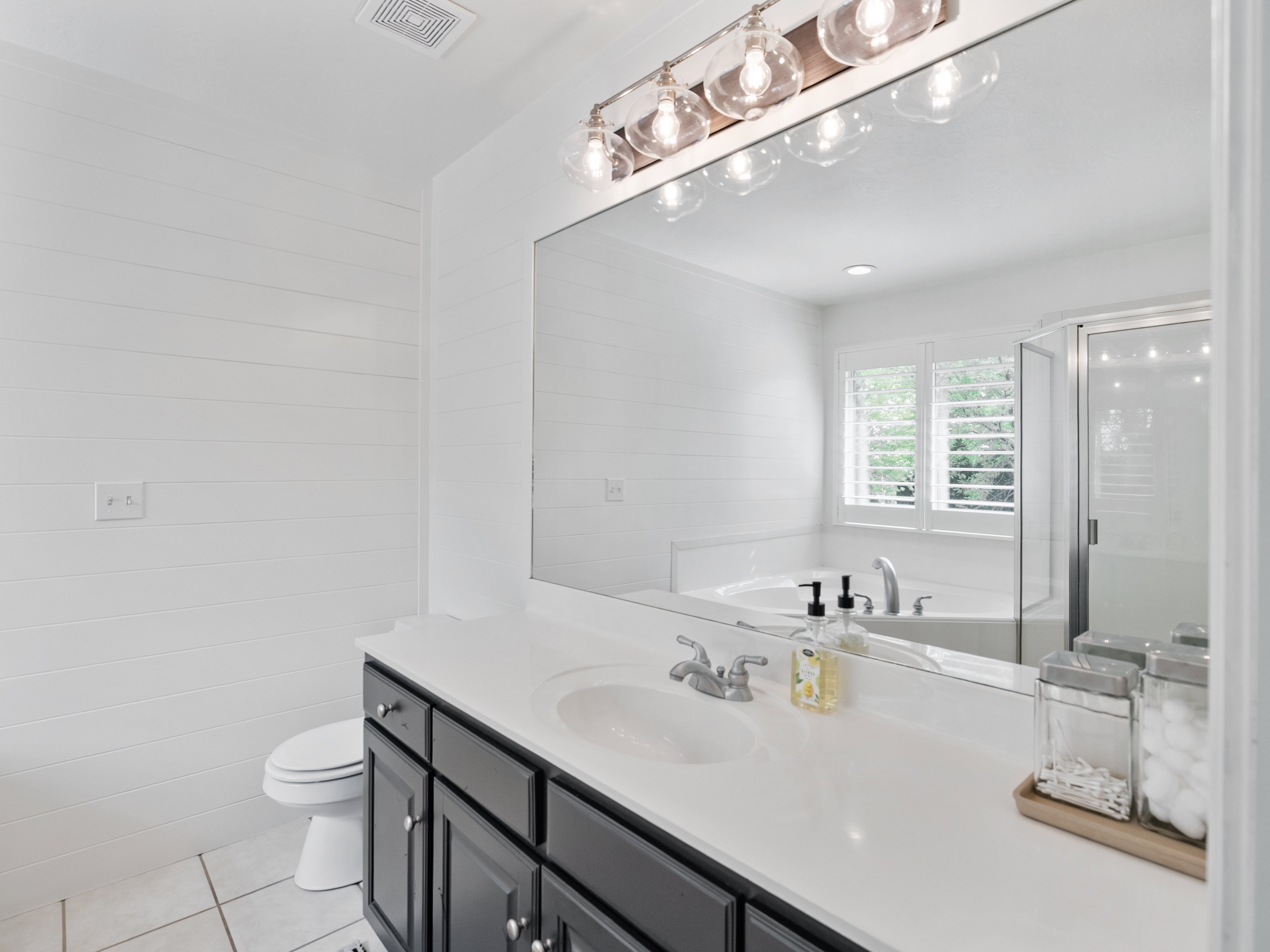 Bathroom featuring a dark wood vanity with a white countertop, large wall mirror, shiplap accent wall, and a garden tub with white plantation shutters