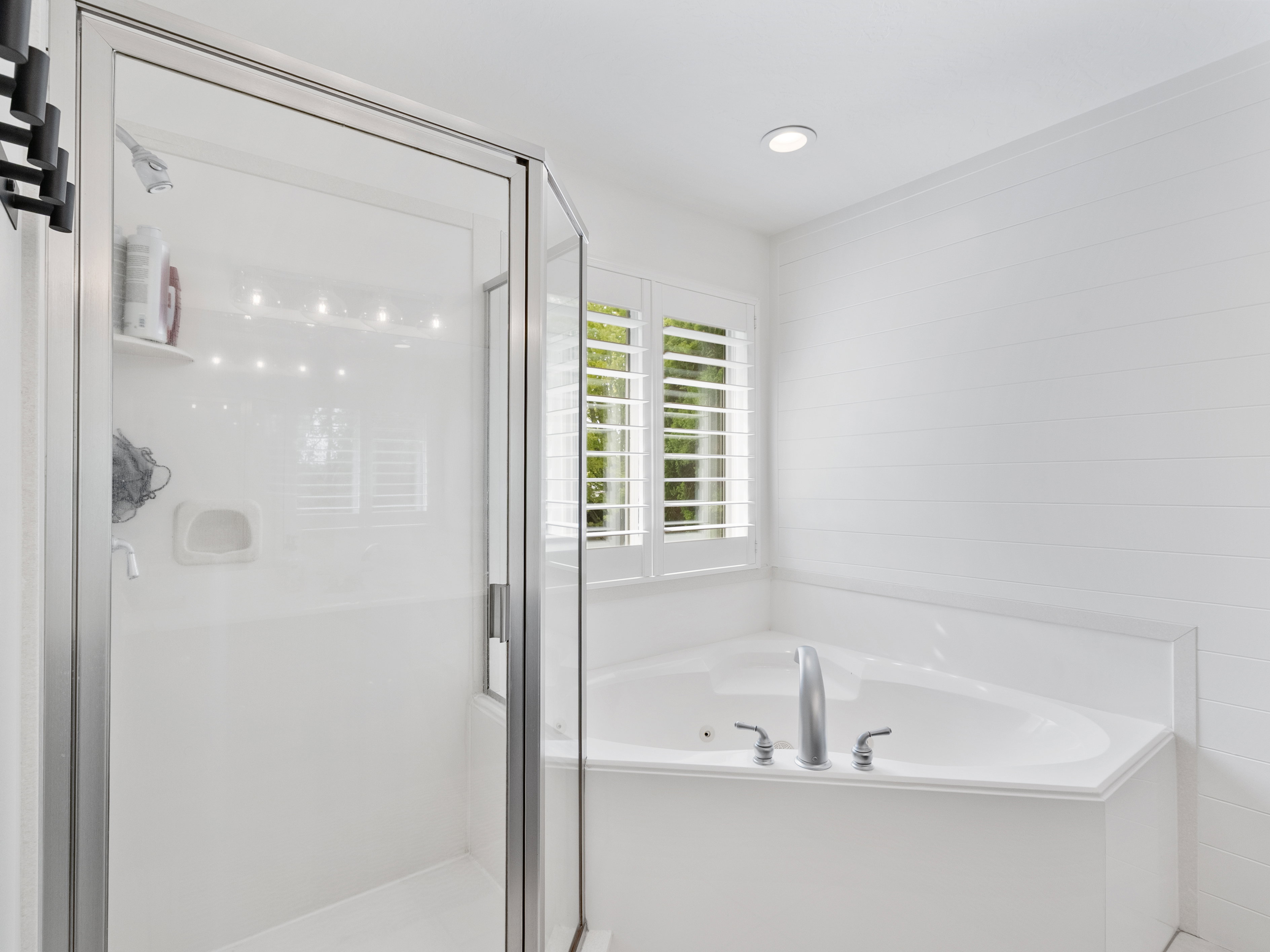Corner soaking tub with deck-mounted fixtures, glass-enclosed shower, white plantation shutters, white horizontal plank wall detailing, and recessed ceiling lighting