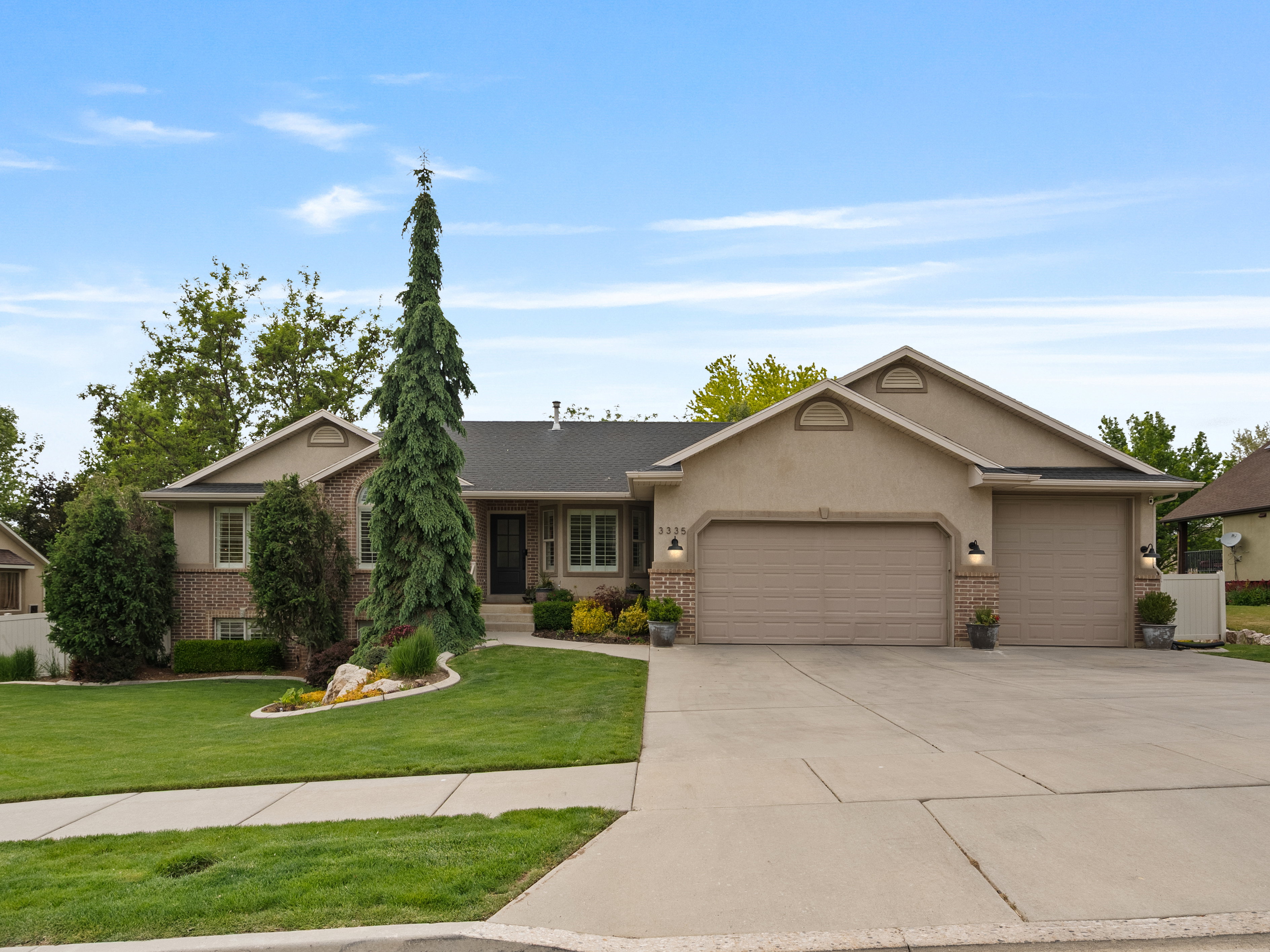Exterior featuring stucco and brick accents, a three-car garage, a concrete driveway, a manicured lawn, and mature landscaping