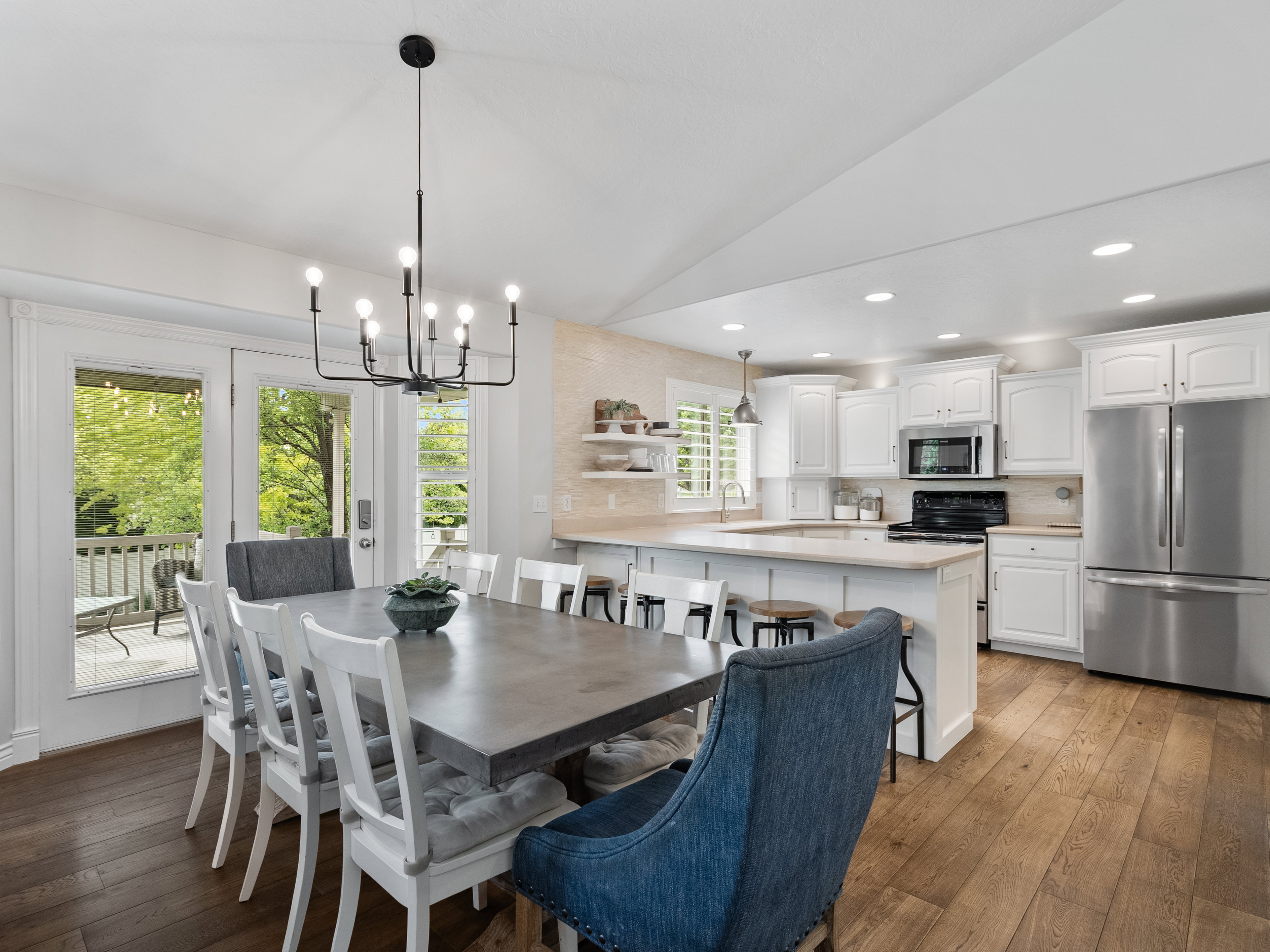 Spacious dining area with a modern black chandelier, wood-finish flooring, and white French doors opening to an outdoor deck