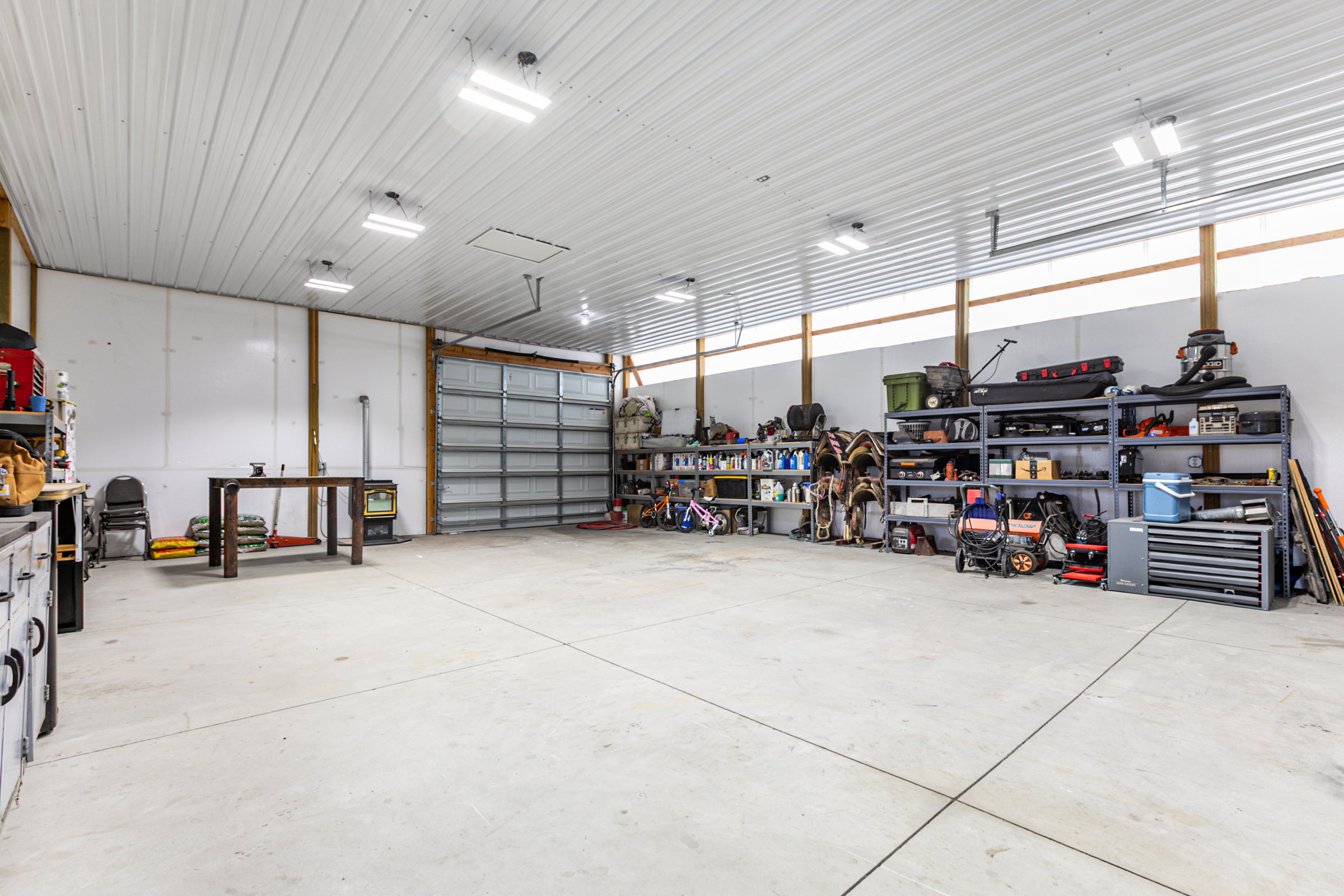 Spacious utility building featuring a concrete floor, high ceilings, exposed wood beams, multiple fluorescent lighting fixtures, and a large roll-up door