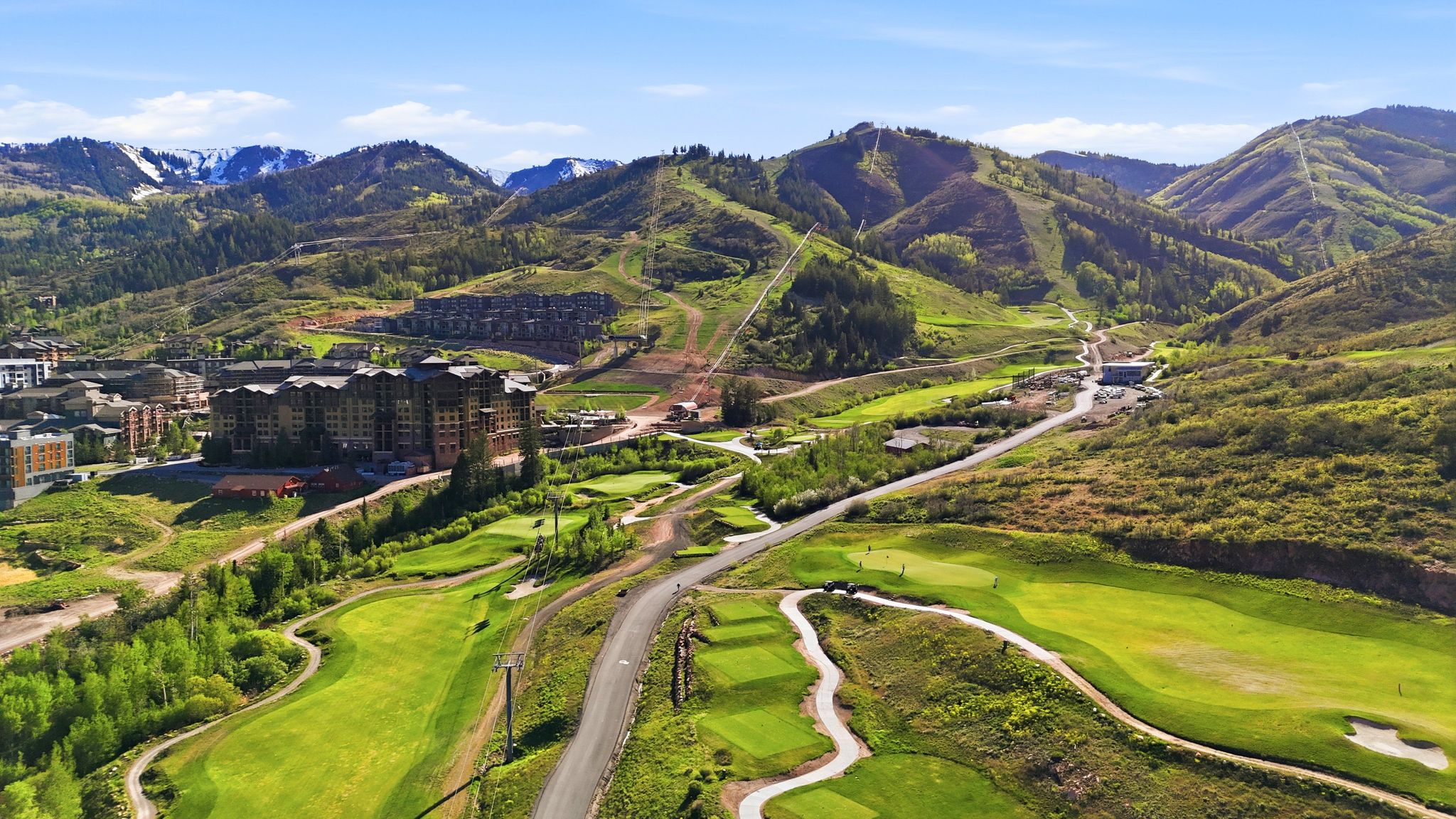 Expansive mountain landscape featuring a golf course, substantial multi-story buildings, and a paved road winding through the terrain