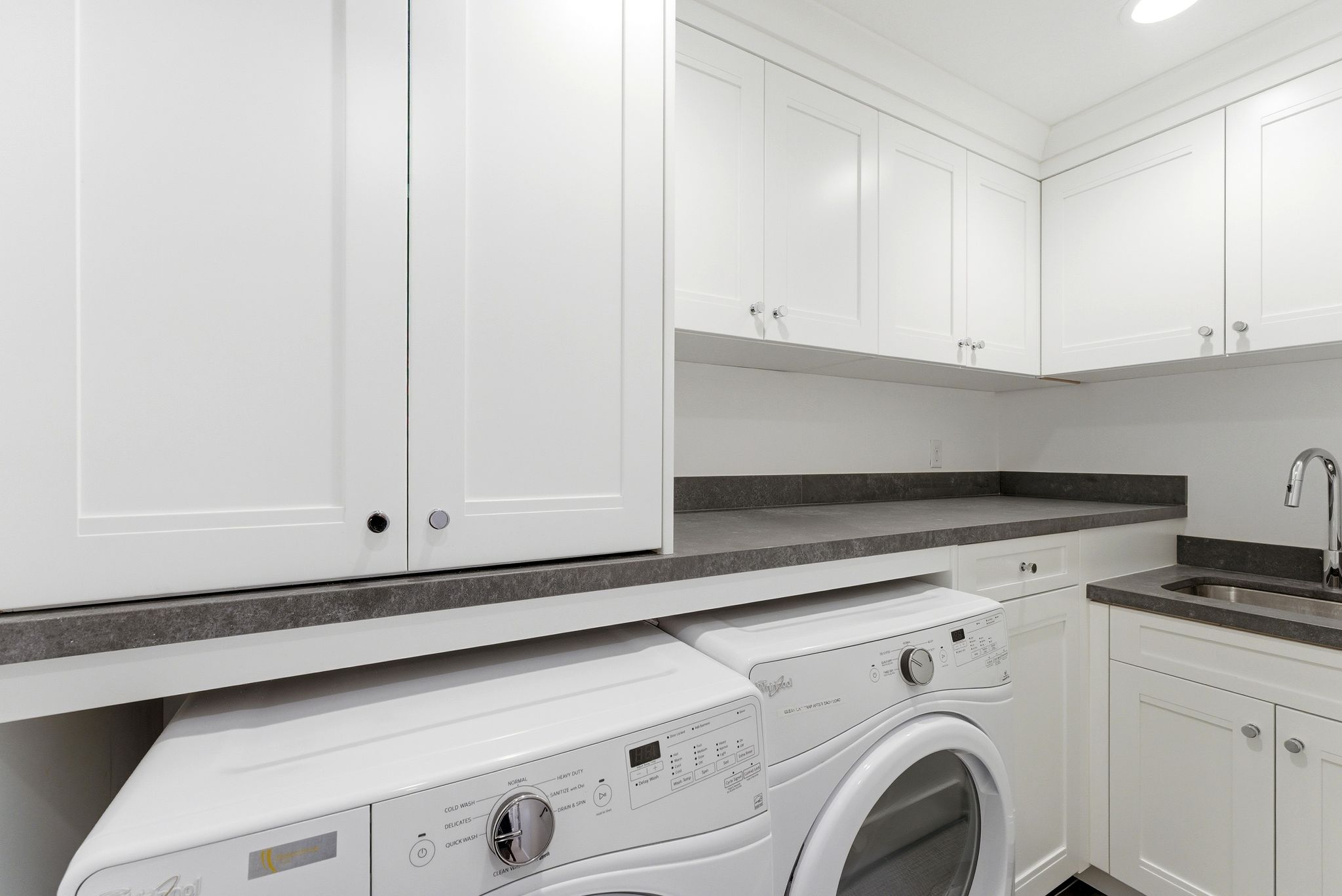 Laundry area featuring white shaker-style cabinetry, dark stone countertops, a stainless steel sink with a chrome faucet, and recessed lighting