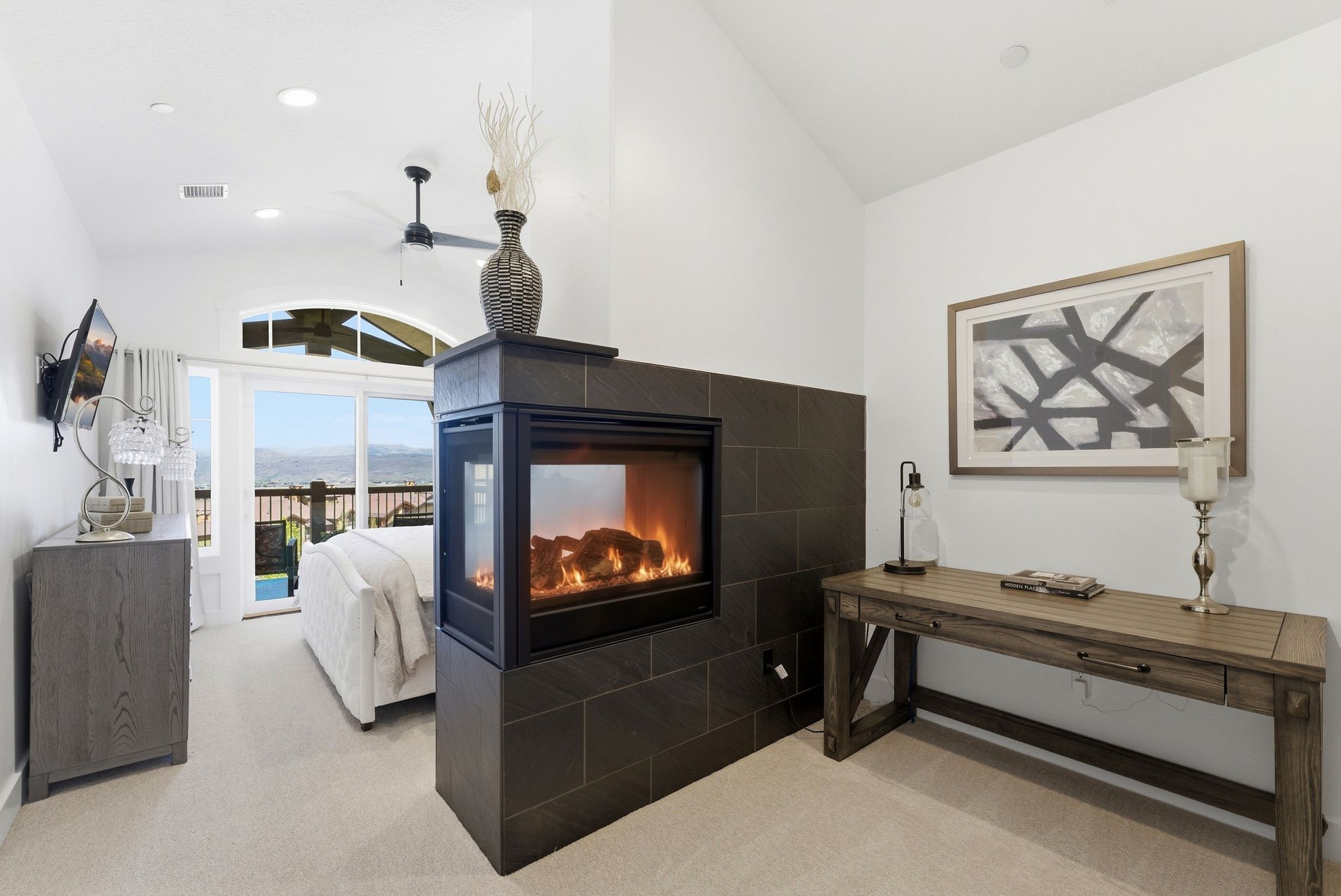 Two-sided fireplace with dark tile surround, vaulted ceiling, recessed lighting, ceiling fan, and sliding glass door leading to a balcony