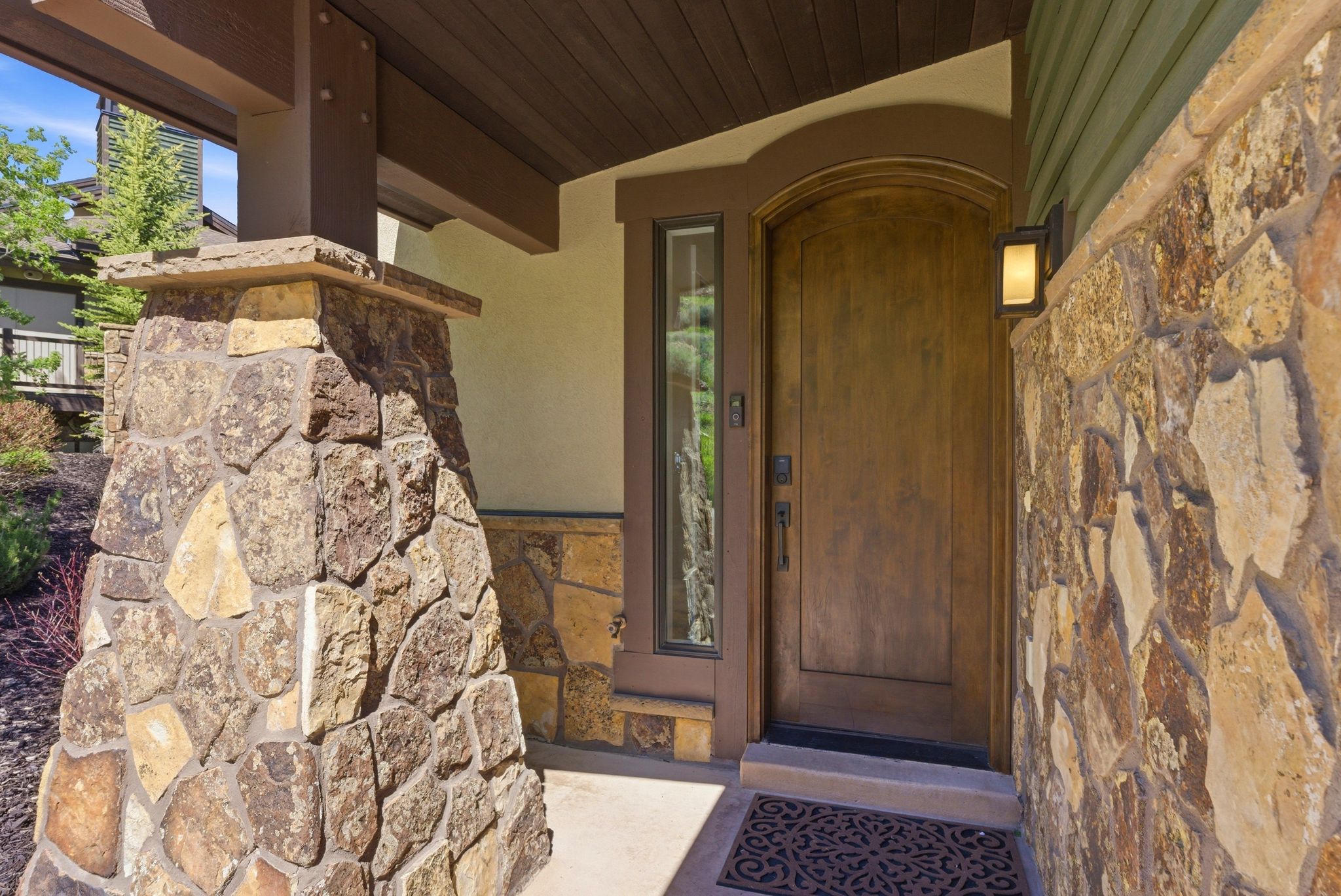 Arched wooden entry door with a vertical sidelight, complemented by stone veneer accents and a wood-paneled soffit