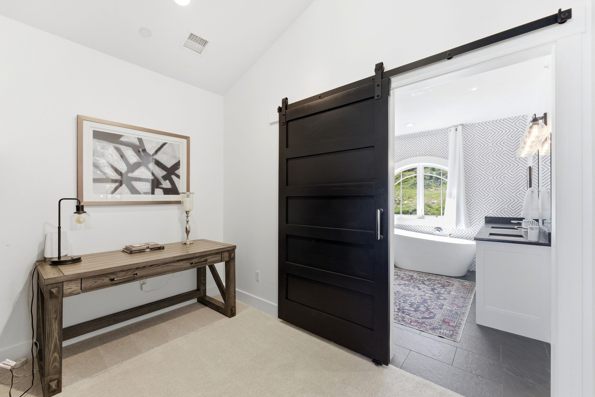 Sliding barn door entry to a bathroom featuring a freestanding soaking tub with a curved window, a single vanity with a dark countertop, and decorative wall tiling