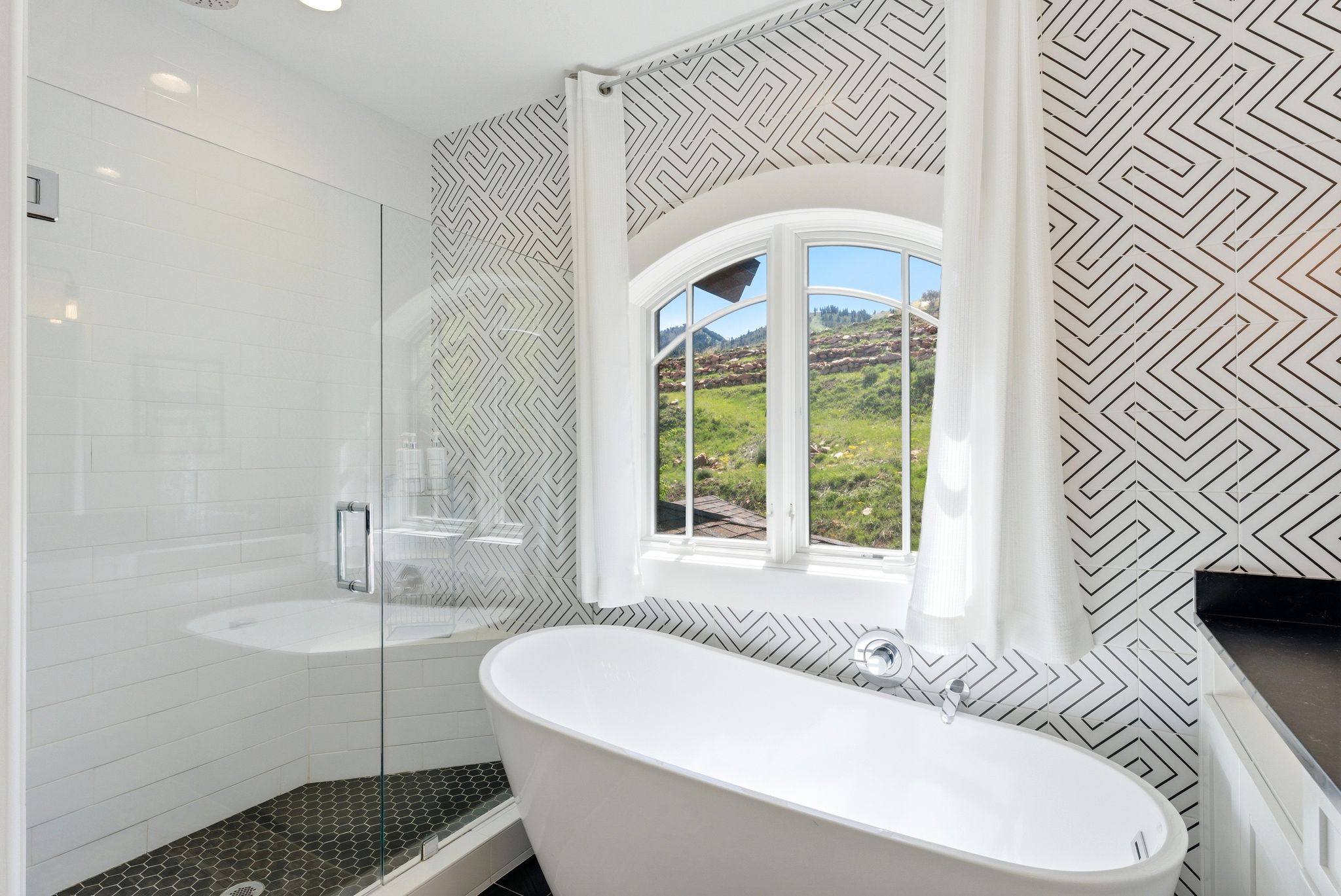 Freestanding soaking tub with arched window, glass-enclosed shower with subway tile, patterned wall covering, and hexagonal floor tile