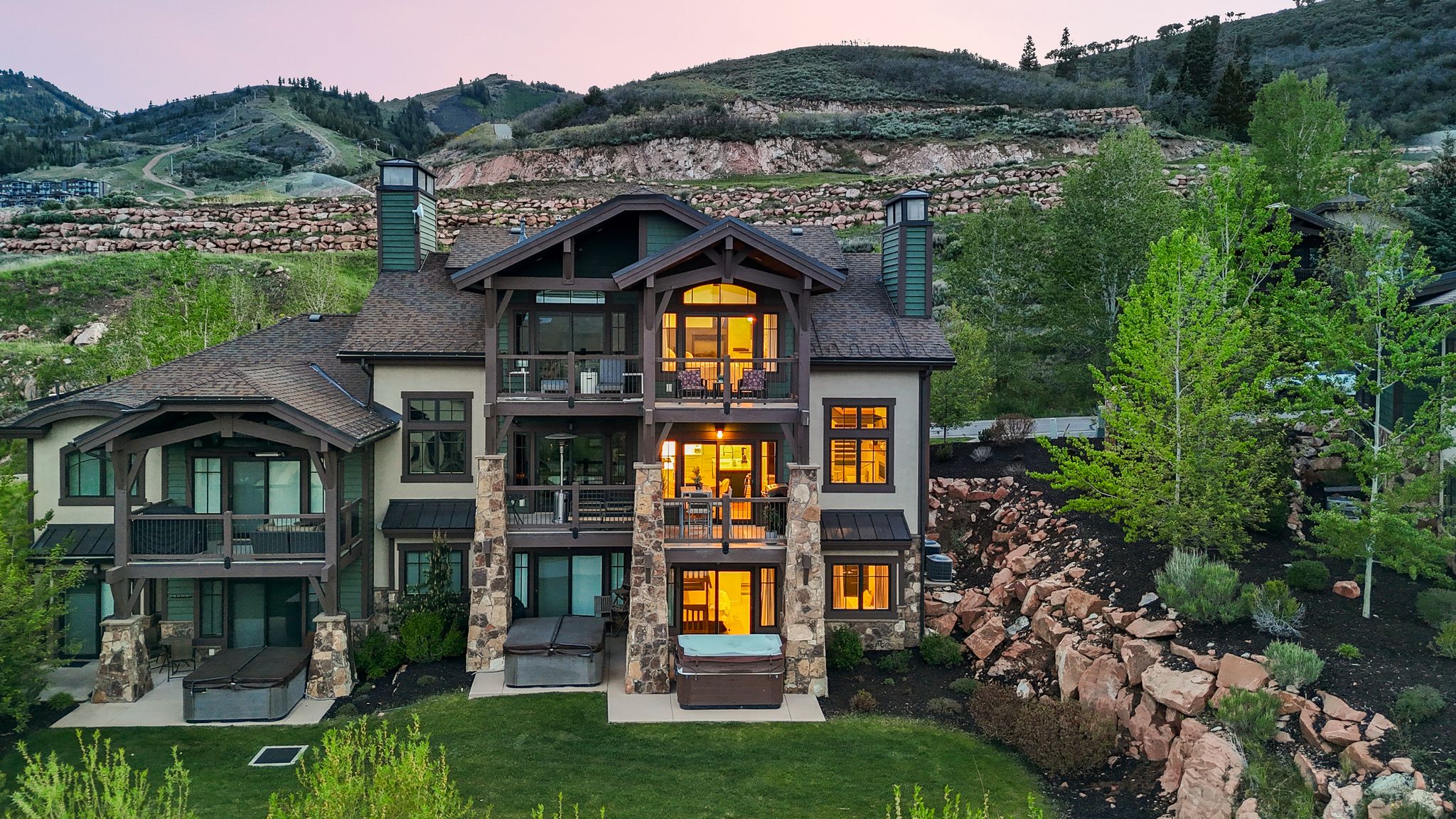 Multi-level mountain modern residence featuring an expansive stone and stucco exterior, multiple covered balconies, a ground-level patio, and three built-in hot tubs
