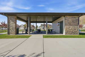 Covered outdoor structure featuring stone-clad pillars, a dark metal roof, concrete flooring, and multiple picnic tables