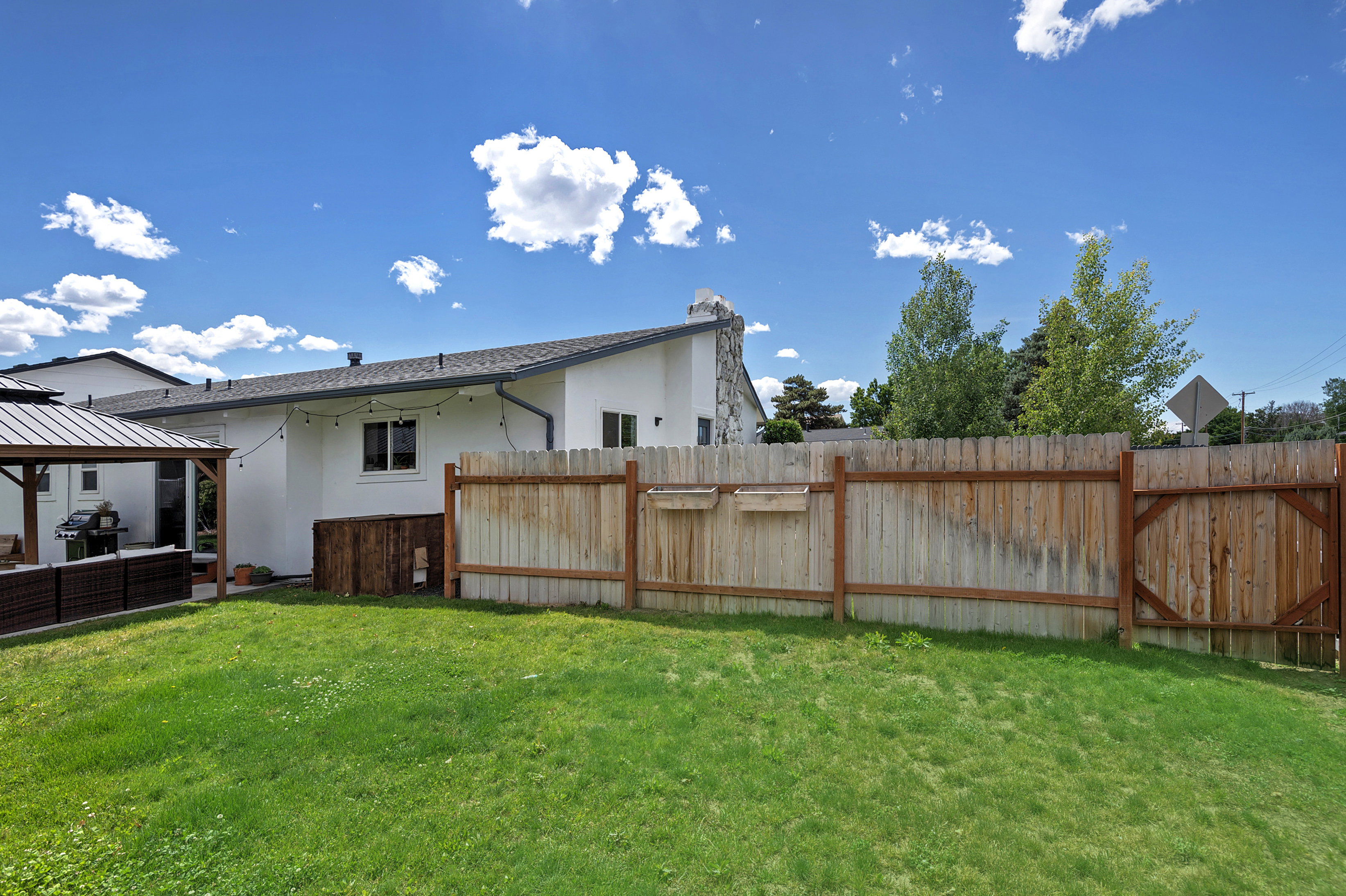Expansive fenced backyard featuring a manicured lawn, a built-in gazebo structure with a metal roof, a white stucco exterior, and a stone chimney