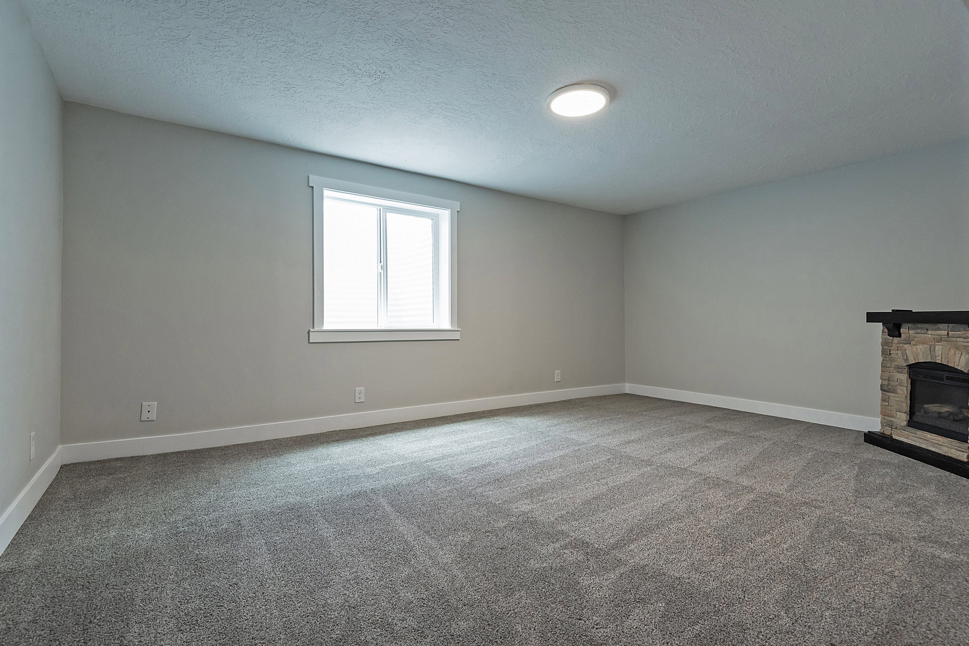 Spacious room featuring gray carpet, a double-hung window with white blinds, and a stone fireplace with a dark mantle