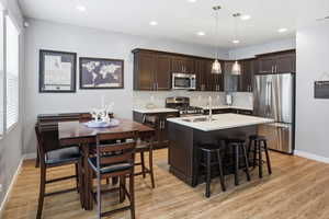 Modern kitchen featuring dark wood cabinetry, stainless steel appliances, a center island with pendant lighting, a tile backsplash, and light wood-finish flooring