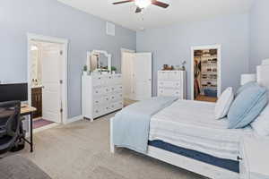 Spacious bedroom featuring light blue walls, soft carpet flooring, an open closet with built-in shelving, a ceiling fan, and a visible doorway to a bathroom with a dark wood vanity and vessel sink
