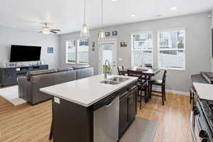 Open-concept living space featuring wood-finish flooring, a kitchen island with a double basin sink and stainless steel dishwasher, a dining area with multiple windows, and a living area with a ceiling fan