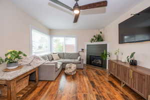 Living room featuring hardwood flooring, corner gas fireplace with a dark paneled surround and white mantel, and a multi-blade ceiling fan with integrated lighting