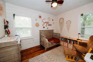 Interior room featuring hardwood flooring, white shiplap accent wall, two windows with blinds, and a ceiling fan with integrated lighting