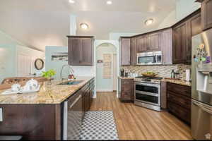 Open-concept kitchen featuring wood-finish flooring, dark wood cabinetry, stainless steel appliances, a multi-tone tile backsplash, and light-toned countertop surfaces