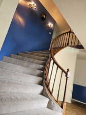 Curved carpeted staircase featuring a wood handrail and balusters, illuminated by two wall-mounted sconces