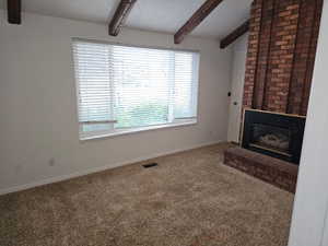 Living area featuring a brick fireplace, exposed wood ceiling beams, a large window with blinds, and neutral-toned carpeting