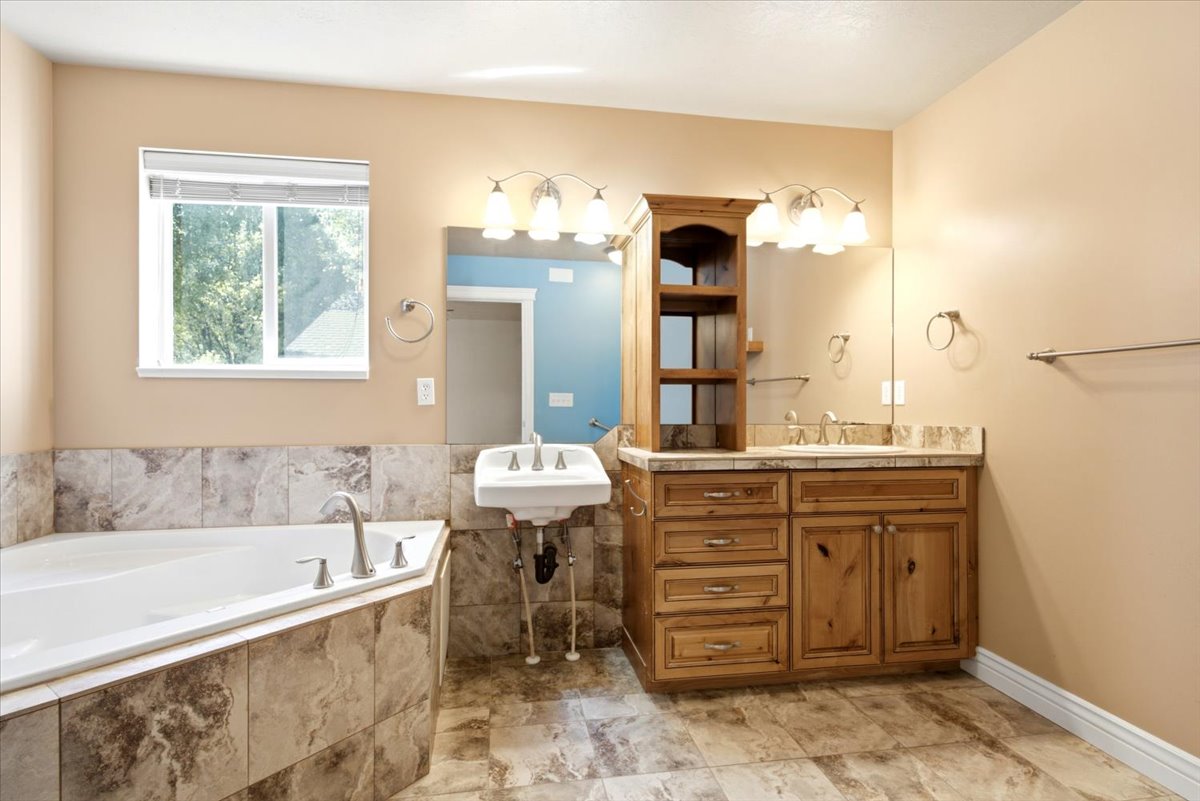 Bathroom featuring a corner soaking tub with tile surround, a double vanity with wood cabinetry and a stone-look countertop, a wall-mounted sink, and tile flooring