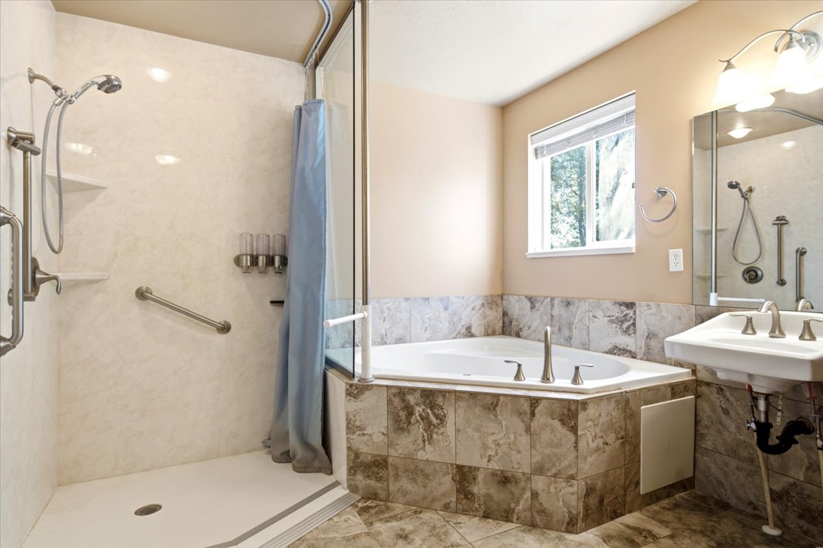 Bathroom featuring a tiled corner soaking tub, walk-in shower with grab bars and built-in shelving, pedestal sink with wall-mounted mirror, and overhead vanity lighting
