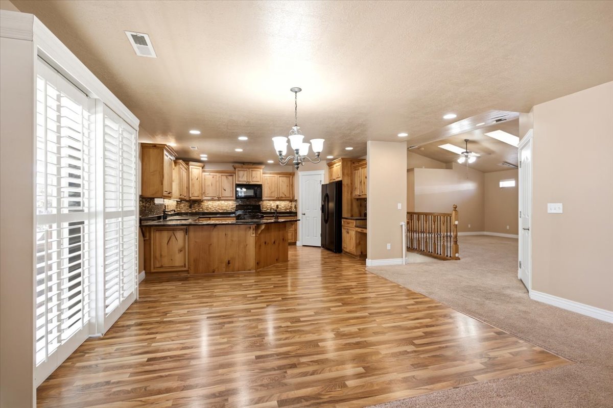 Open-concept living space featuring wood-finish flooring, a kitchen with wood cabinetry and dark countertops, recessed lighting, and white plantation shutters