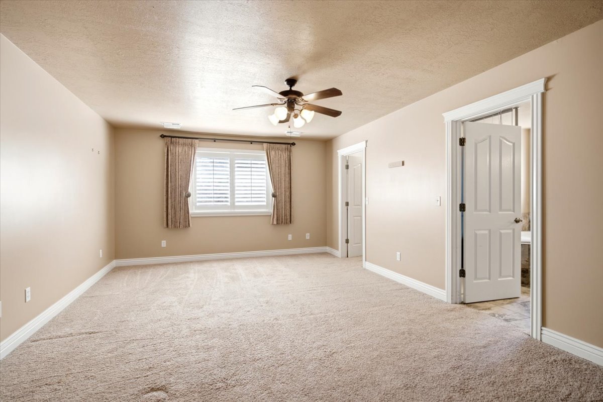 Spacious room featuring neutral carpeting, a ceiling fan with integrated lighting, and a window with white louvered shutters and drapes