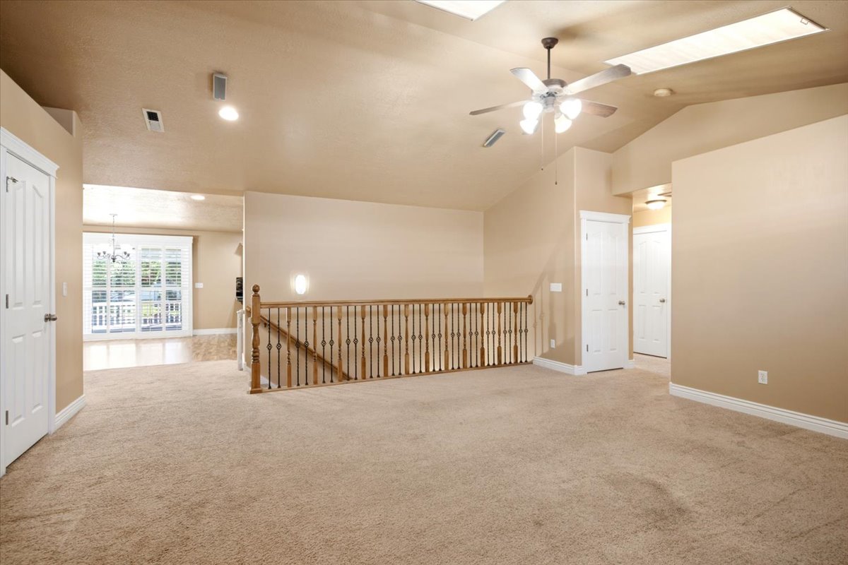 Spacious carpeted area featuring a wood staircase with iron balusters, vaulted ceilings with skylights, and a ceiling fan with integrated lighting