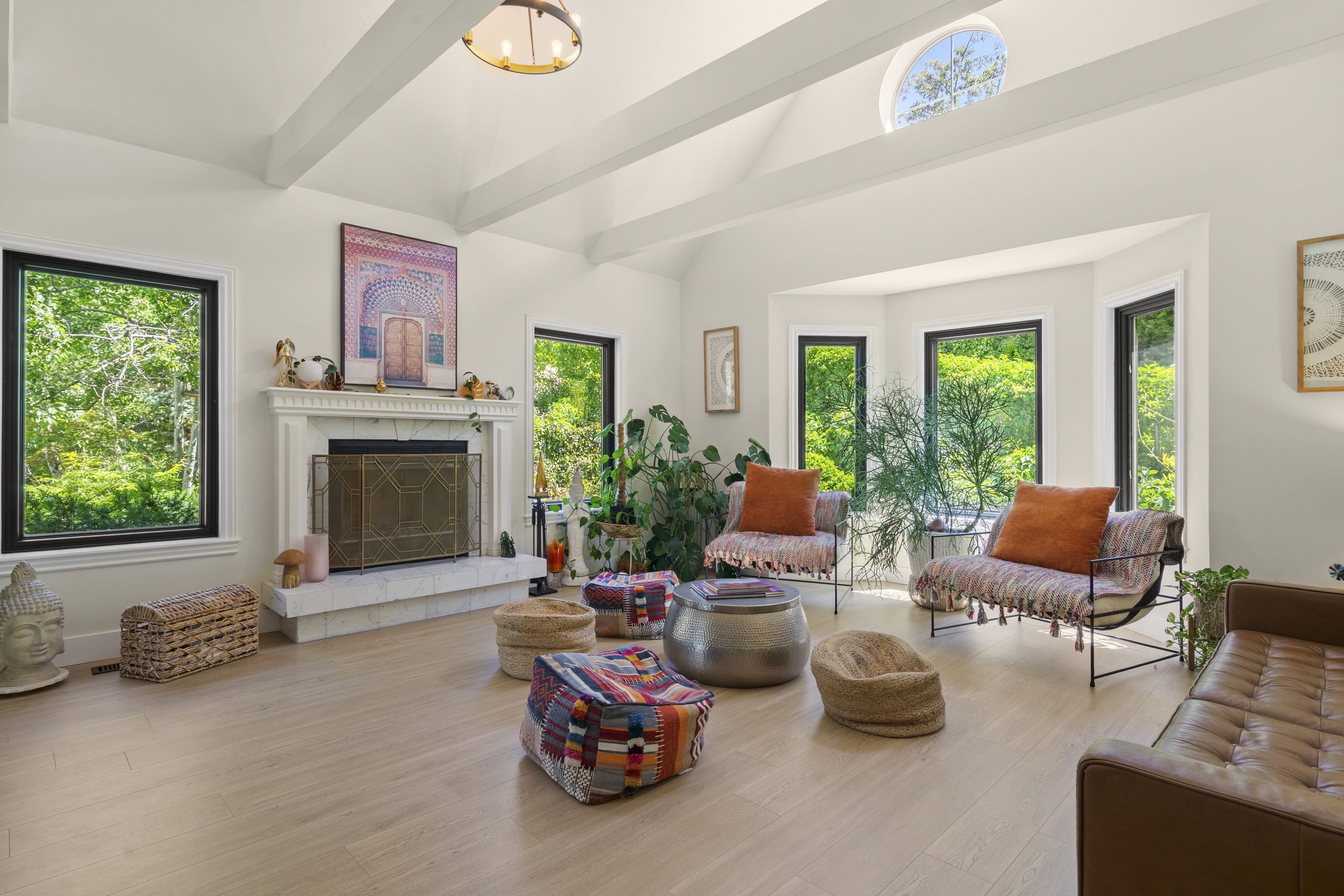 Spacious interior featuring a white fireplace with a decorative screen and mantel, vaulted ceilings with exposed beams, light wood-finish flooring, a bay window configuration, and a prominent circular window